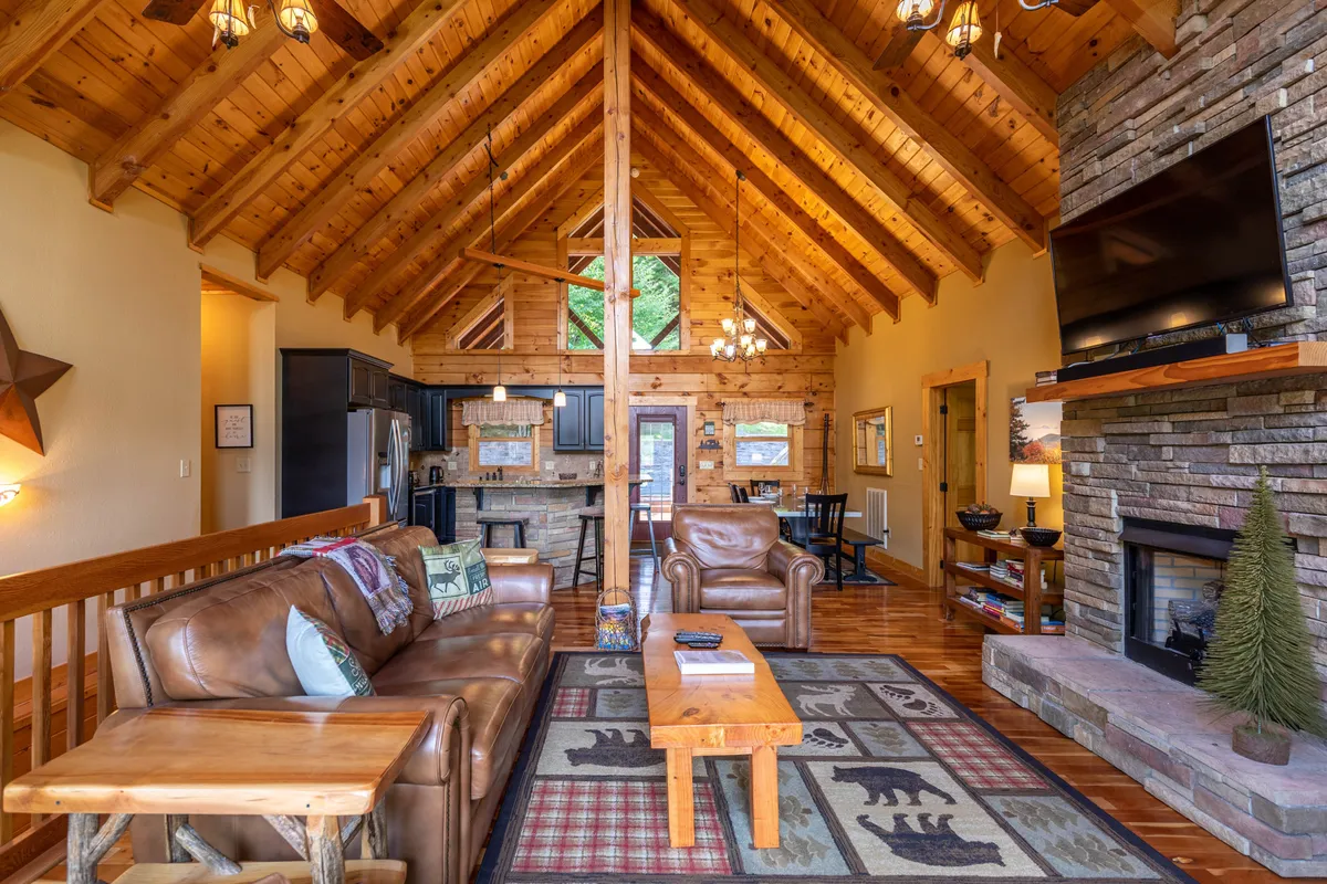 A warm and welcoming interior living space featuring a leather sofa, stone fireplace, wooden beams, and a rug with bear motifs. The area is open and cozy, ideal for relaxing after a day outdoors.