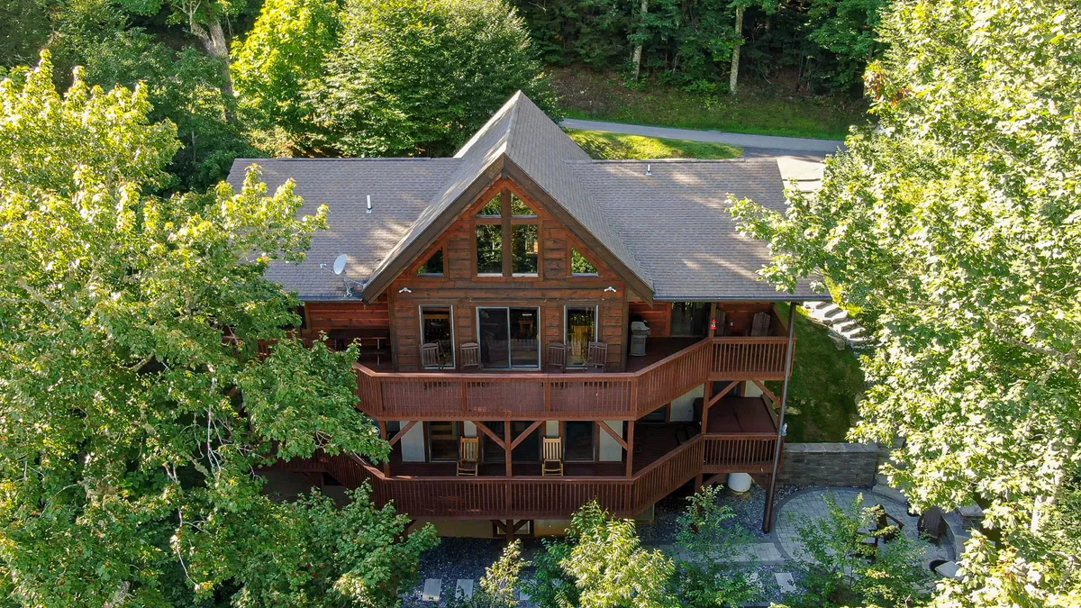 The image showcases the cabin's striking wooden architecture set amidst lush greenery, with expansive windows and inviting balconies overlooking the scenic Sugar Mountain landscape. The balconies offer cozy seating, perfect for relaxing and taking in sunset views or stargazing at night.