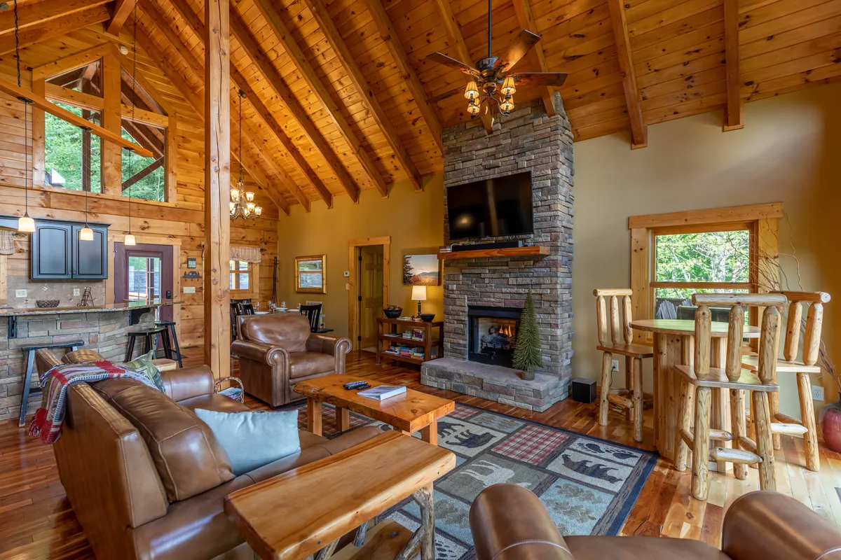 An open living and dining area with high wooden ceilings, a stone fireplace, comfortable sofas, and a spacious dining table positioned near large windows letting in abundant natural light.