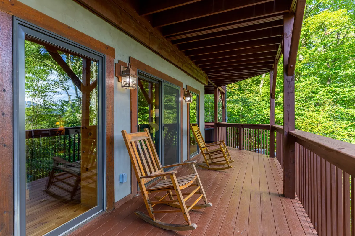 An inviting exterior deck with wooden rocking chairs and large windows, surrounded by lush greenery. The tranquil setting suggests a place to unwind with a loved one while soaking in the views and fresh mountain air.