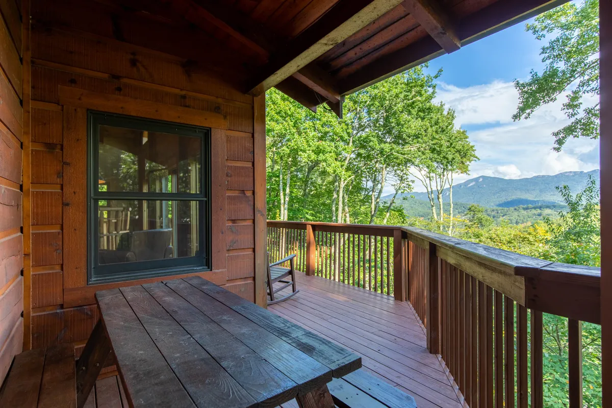 An inviting outdoor table set on a deck overlooking sweeping mountain vistas, bordered by green trees. The peaceful setting encourages group gatherings, reading, or enjoying sunrise and sunset over the mountains.