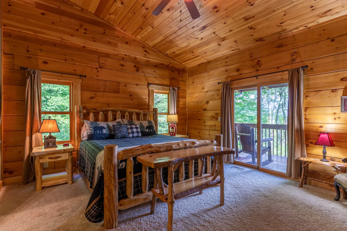A bright, welcoming bedroom featuring a rustic log bed, large windows that let in an abundance of natural light, and cozy bedside lamps. The room exudes calm and relaxation, with views of the outdoors and a plush rug underfoot.