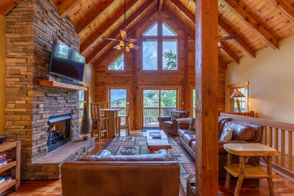 A cozy great room with a striking stone fireplace, exposed wooden beams, and expansive windows framing breathtaking mountain views. Plush seating invites relaxation while the architectural details highlight the soaring ceilings.