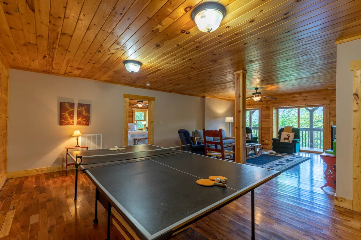 The image captures a ping pong table set beneath a wooden ceiling, with large windows providing exterior views and a comfortable seating area close by. The scene is bright and inviting, suggesting a space where both play and relaxation happen side by side.