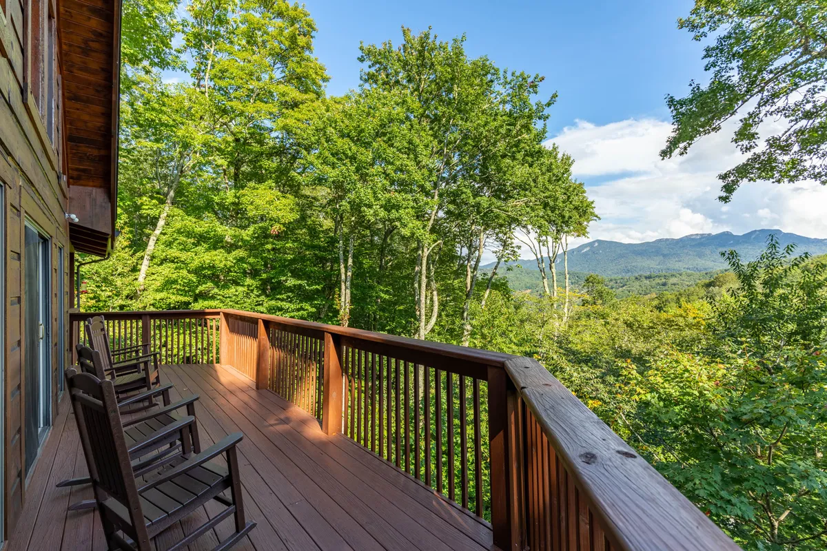 A peaceful wooden deck overlooks sweeping mountain vistas, framed by lush greenery. Rocking chairs invite guests to relax and soak in the view, creating an idyllic spot for morning coffee, sunset cocktails, or quiet stargazing. The deck is spacious and seamlessly blends with the surrounding landscape, embodying the essence of indoor/outdoor mountain living.