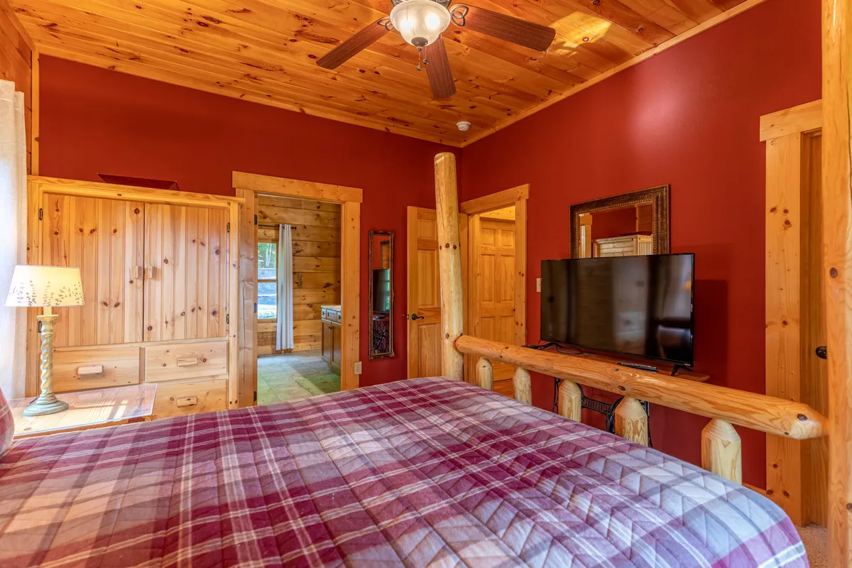 A bedroom with a plaid bedspread, wooden furniture, a television mounted on the wall, and a lamp on the nightstand. The room is warm, inviting, and suitable for relaxation or social moments.