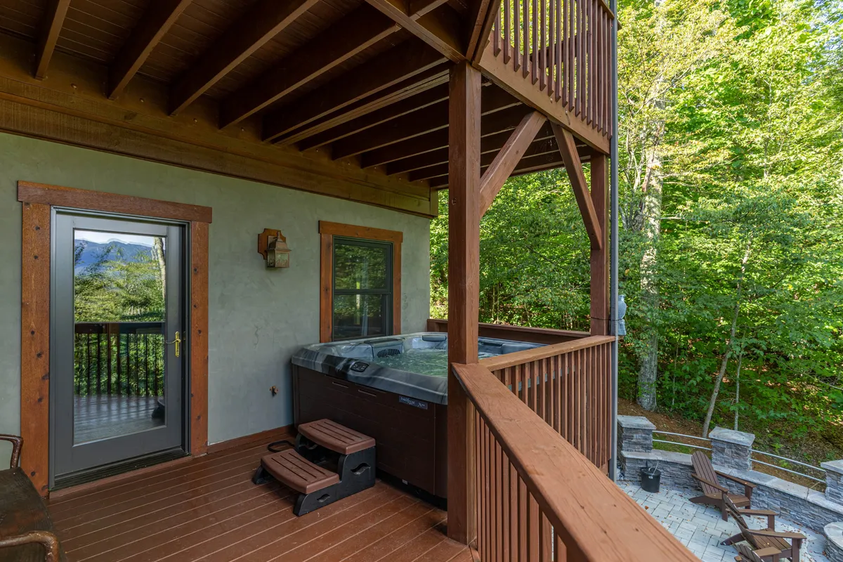 A tranquil outdoor scene featuring a hot tub on a wooden deck, surrounded by lush trees. The setting offers expansive views of nature, making it ideal for unwinding after an adventurous day. The spacious deck also hints at room for group gatherings and relaxation under the open sky.
