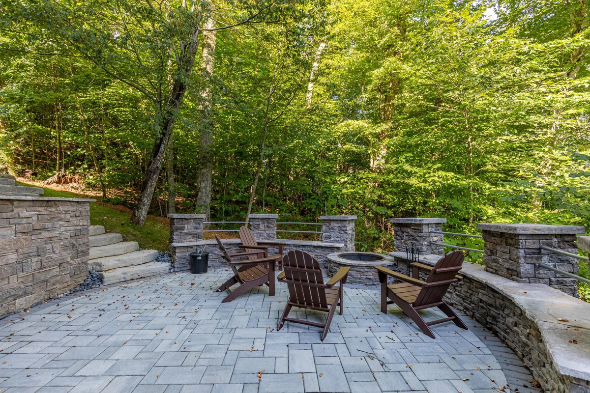 A stone patio nestled among the trees features a fire pit surrounded by Adirondack chairs. The inviting setup suggests cozy evenings spent with loved ones, sharing stories, roasting marshmallows, or simply gazing at starry skies. The fire pit and natural setting embody the convivial, outdoorsy spirit of the home.